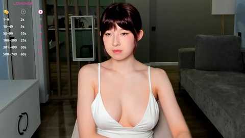 Media: A video of a young Asian woman with short dark hair, fair skin, and large breasts in a low-cut white tank top, sitting in a modern living room with a grey sofa and wooden floor.