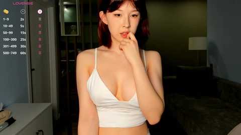 Media: Video of an East Asian woman with fair skin, red hair, and bangs, wearing a white, low-cut tank top, biting her finger, indoors with dim lighting, and a modern, minimalist background.