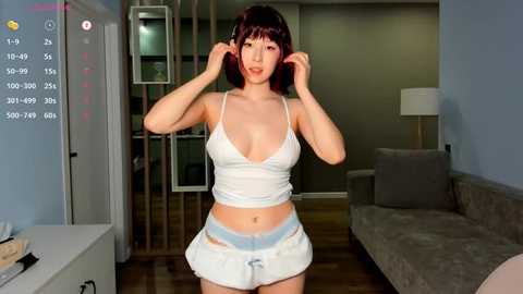 Media: Video of an Asian woman with short brown hair, wearing a revealing white tank top and ripped denim shorts, standing in a modern living room with light walls and a grey sofa.