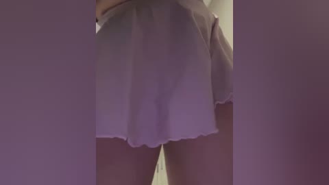 Media: Video of a woman's lower body, showing her light-colored, frilled short skirt, highlighting her bare thighs. She stands in a dimly lit room with muted purple walls, suggesting an intimate or private setting.