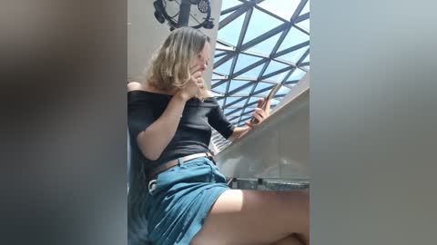 Media: Video of a blonde woman with shoulder-length hair, wearing a black off-the-shoulder top and teal shorts, seated near a window with a geometric patterned glass, holding her hand to her mouth, looking thoughtful.