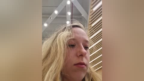 Media: Video of a blonde woman with messy hair, looking serious, in a room with beige walls, wooden slats, and white ceiling lights.