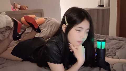 Media: A video of an Asian woman with long black hair, wearing a black dress and red high heels, lying on a bed with a green lighted sex toy beside her.