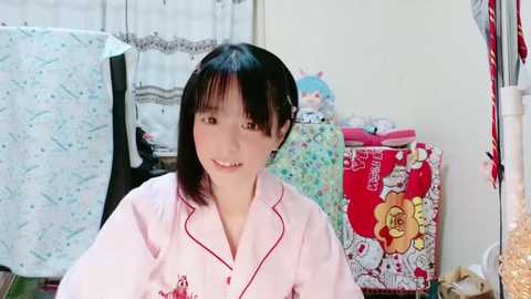 Media: Video of a young East Asian woman with straight black hair, smiling in a pink pajama top, surrounded by colorful, patterned fabric in a cluttered bedroom.