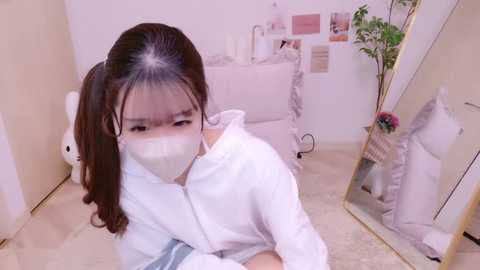 Media: Video of an Asian woman with long brown hair in a white mask, wearing a white bathrobe, sitting in a beige tiled room with a mirror, potted plant, and white furniture.