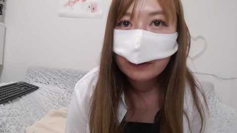 Media: Video of a young woman with long, straight brown hair, wearing a white surgical mask, a black top, and a white lab coat. She is indoors, seated on a bed with gray bedding, in a sterile-looking room.