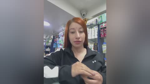 Video of a young, fair-skinned woman with long, straight red hair, wearing a black Adidas jacket, adjusting her hair in a dimly lit, cluttered store with shelves full of products.