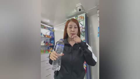 Video of a young Asian woman with long red hair, wearing a black jacket, holding a water bottle in a modern, brightly lit convenience store.