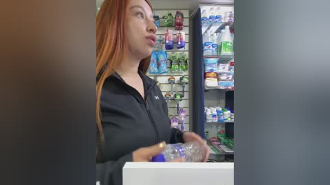 Media: Video of a young redhead woman in a black jacket, holding a water bottle in a pharmacy. Background features shelves with various products, including skincare and medical supplies.