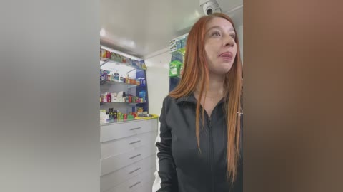 Video of a red-haired woman with long hair, wearing a black jacket, standing in a brightly lit pharmacy with colorful shelves filled with various products.