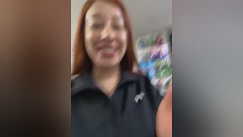 Media: A blurred video of a smiling woman with light brown skin and long auburn hair, wearing a black polo shirt. Background features colorful, abstract art.