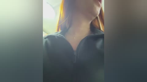 Video of a fair-skinned woman with long, straight red hair, wearing a dark, zippered hoodie, taken indoors with blurred background and soft lighting.