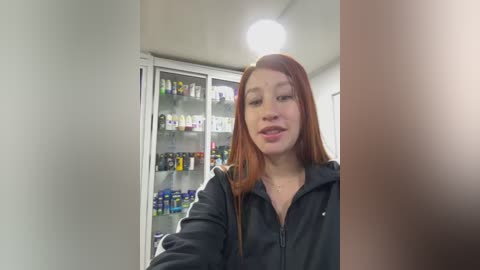 Video of a young woman with long, straight red hair, wearing a black jacket, standing in a brightly lit pharmacy with shelves full of various products in the background.