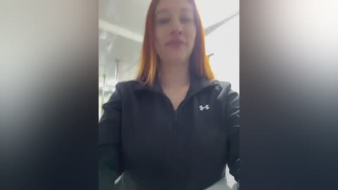 Video of a young Caucasian woman with straight, shoulder-length red hair, wearing a black Under Armour zip-up hoodie and gray pants, standing indoors with blurred background.