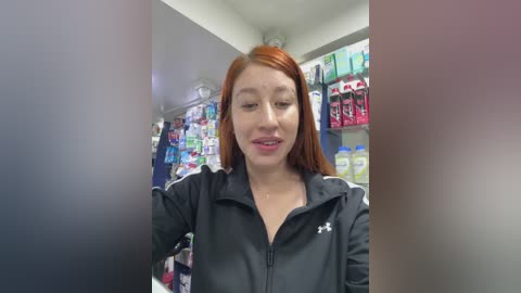 Video of a young woman with fair skin and long red hair, wearing a black zip-up jacket with white stripes. She is standing in a brightly lit convenience store with shelves filled with various products.