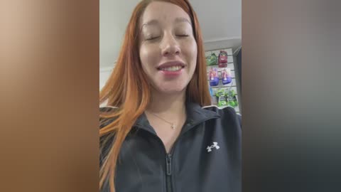 Media: A candid video of a smiling young woman with long, straight, red hair, wearing a black Under Armour jacket, standing in a well-lit room with shelves of colorful products in the background.