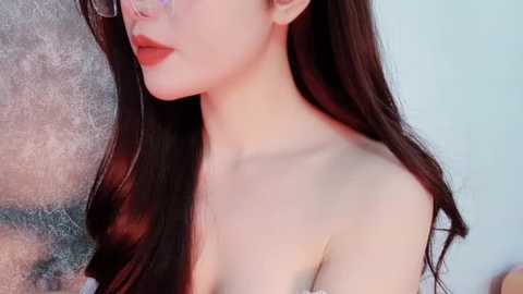 Media: Video of a fair-skinned, East Asian woman with long, dark hair and red lipstick, seen from the side. She's topless, with a textured wall in the background.