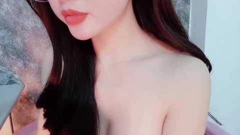 Media: A video of a young East Asian woman with fair skin, long black hair, and full red lips. She is topless, with her left shoulder and collarbone visible. The background is a blurred, textured, gray wall.