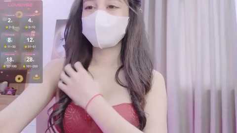 Media: A video of an Asian woman with long black hair, wearing a red lace bra and white mask, adjusting her hair in a bedroom with a pink and white curtain background.