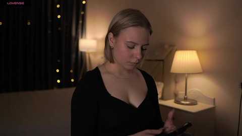 Media: Video of a blonde woman in a black top, reading a smartphone in a dimly lit room with a white lamp and dark curtains.