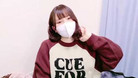 Video of an Asian woman with straight, shoulder-length brown hair, wearing a white face mask and a maroon and cream sweatshirt with \"CUED\" in bold letters. She is indoors with light-colored walls and a curtain in the background.