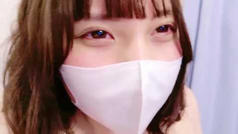 Video of an East Asian woman with shoulder-length brown hair, wearing a white surgical mask covering her nose and mouth, and light blue shirt.