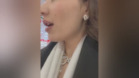 Media: A close-up video of a woman's profile, wearing a black blazer over a white shirt, with a silver necklace and earrings. The background is blurred, with indistinct red and white signage.