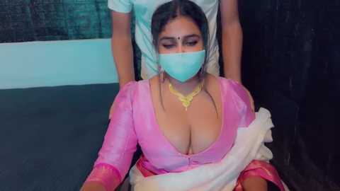 Media: Video of a South Asian woman with a light brown complexion, wearing a pink sari, a turquoise mask, and gold jewelry, restrained in a chair by a man, in a dimly lit room.