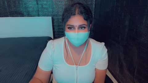 Media: Video of a South Asian woman with medium skin tone, wearing a white short-sleeved top and a blue surgical mask, sitting on a black bed with a white headboard, against a dark brick wall background.