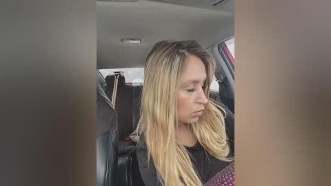 Media: Video of a blonde woman with long hair in a black top, sitting in a car with grey and black interior, looking sad.