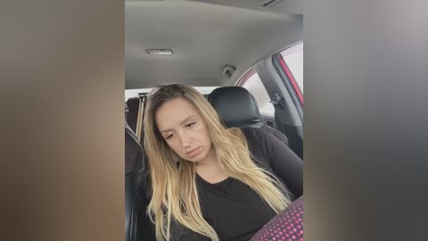 Media: Video of a young woman with long, wavy blonde hair, wearing a black top, sitting in a car, looking tired and slightly pouting, with blurred car interior in the background.