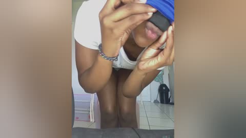 Media: Video of a dark-skinned woman with medium-sized breasts, wearing a white t-shirt and shorts, holding her head with hands, in a tiled bathroom.