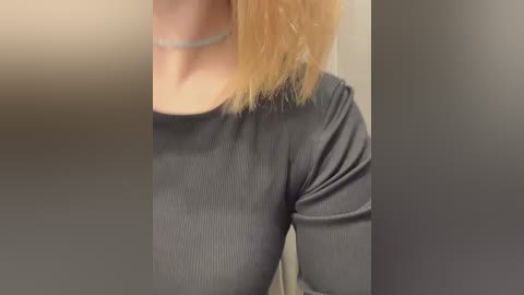 Media: A video of a woman with fair skin and shoulder-length blonde hair, wearing a tight, ribbed, dark grey top. She has a thin, silver choker necklace. The background is blurred, likely a bathroom mirror.