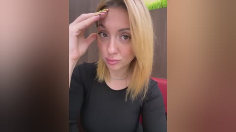 Media: Video of a young Caucasian woman with blonde hair, wearing a black long-sleeved top, touching her forehead. Background is blurred with brown and red hues.