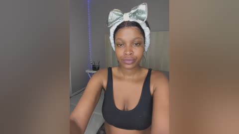 Media: Video of a young Black woman with medium skin tone, wearing a black sports bra, white headband with a large green bow, and a neutral expression. The background shows a simple, modern room with light walls and a white door.