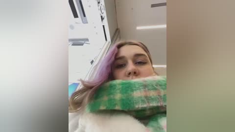 Media: Video of a young Caucasian woman with blonde hair, partially covered by a green and white plaid scarf, in a white subway car.