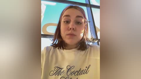 Media: A video of a young woman with straight, shoulder-length auburn hair, wearing a white t-shirt with \"The Cocktail Hour\" printed in cursive. She has a neutral expression. Background features a window with a blue sky and white clouds.