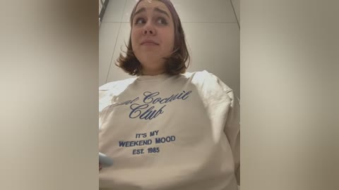 Media: Video of a young woman with short brown hair, wearing a beige T-shirt with \"The Orchid Club\" and \"It's my weekend wood, est. 2004\" printed in navy blue. She has a neutral expression, standing in a tiled bathroom.