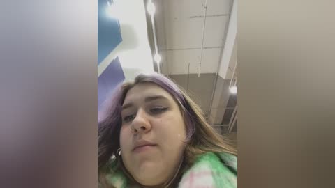 Media: Video of a young woman with light skin, brown hair, and a green tie-dye shirt, sitting in a dimly lit bathroom with white tiles, visible mirror, and blue towels.