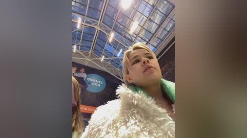 Media: Video of Miley Cyrus, a young Caucasian woman with blonde hair, wearing a white fur coat, standing in an indoor venue with a glass ceiling and promotional posters in the background.