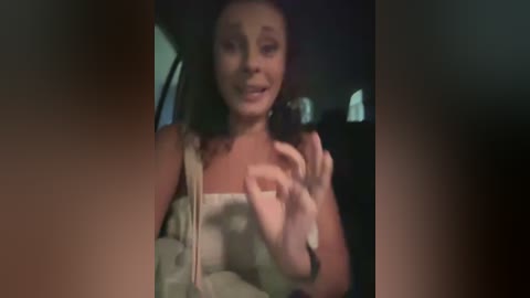 Video of a woman with light skin and dark hair, wearing a white tank top, smiling and holding a phone, in a dimly lit car interior.