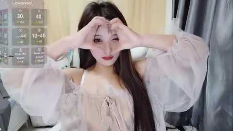 Media: Video of an Asian woman with long black hair, fair skin, and red lipstick, wearing a sheer, off-shoulder top, making a heart shape with her fingers against a backdrop of curtains and a digital screen displaying health data.