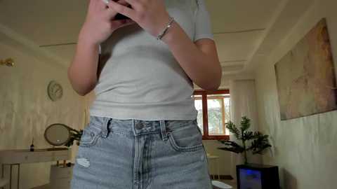 Video of a woman in a white t-shirt and high-waisted, distressed denim shorts, standing in a cozy, beige-walled room with a window, potted plant, and abstract painting in the background.