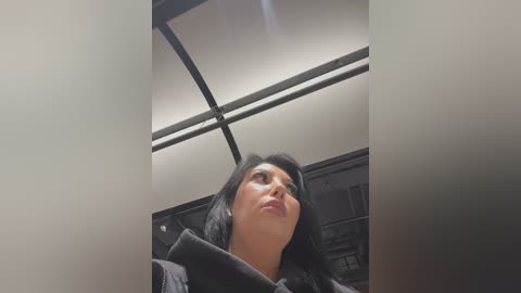 Media: Video of a woman with long black hair and light skin, wearing a black jacket, looking up in an indoor setting with a metal ceiling and industrial elements.