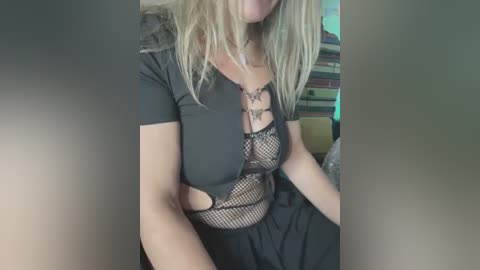 Media: Video of a blonde woman with fair skin wearing a black, sheer, fishnet top revealing her medium-sized breasts, and a black skirt, seated in a room with green and yellow furniture.