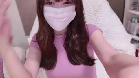 Media: Video of a woman with long brown hair, wearing a white face mask, lavender shirt, and taking a selfie in a bedroom with a white bed and nightstand.