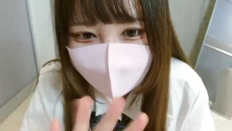 Media: Video of an Asian woman with long brown hair, wearing a white surgical mask, brown eyes, and a white lab coat, indoors with a light-colored background.