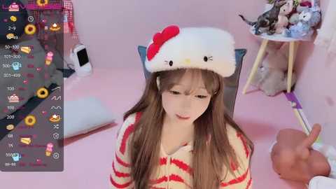 Video of a young woman with long brown hair, wearing a striped sweater and a white Hello Kitty hat, sitting on a chair. Background includes a pink room with plush toys and a phone showing various icons.