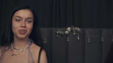 Video of a young woman with long black hair, wearing a sleeveless dress, and pearl necklace, speaking against a dark curtain backdrop adorned with floral arrangements and jewelry displays.