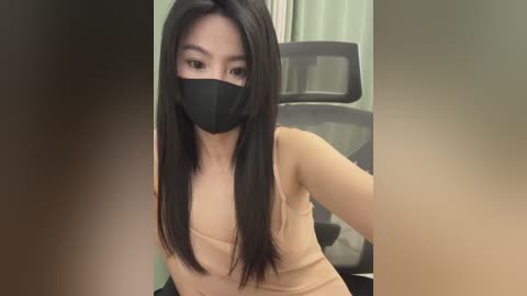 Video of an Asian woman with long black hair, wearing a black face mask, beige tank top, and sitting in an office chair. Background includes a blurred, beige wall.